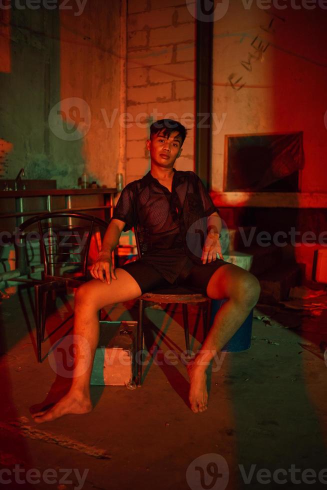 un gay hombre vistiendo negro y posando con un silla en un antiguo casa con un rojo ligero foto