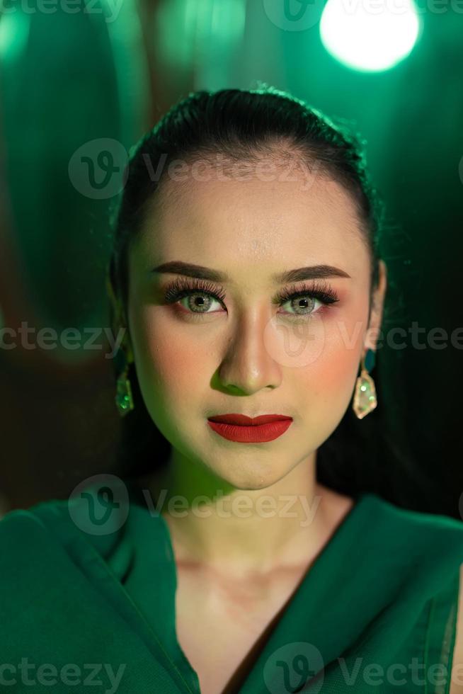 cerca arriba foto de un asiático mujer con un lleno cara de maquillaje y vistiendo verde ropa y joyería en su orejas
