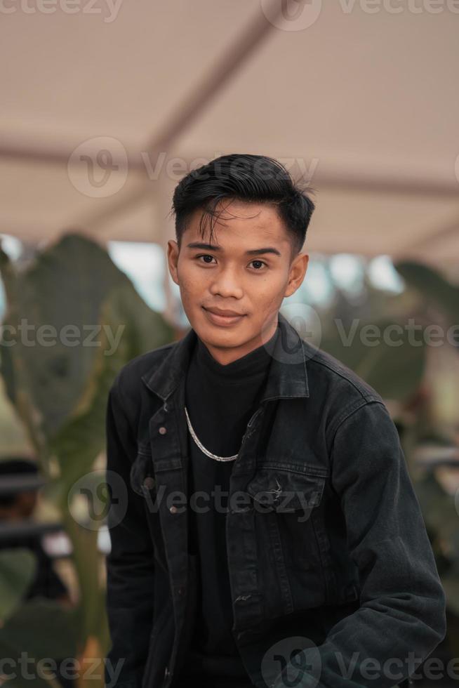 an Asian man in a black denim jacket and a chain necklace posing in a relaxed style in a park photo