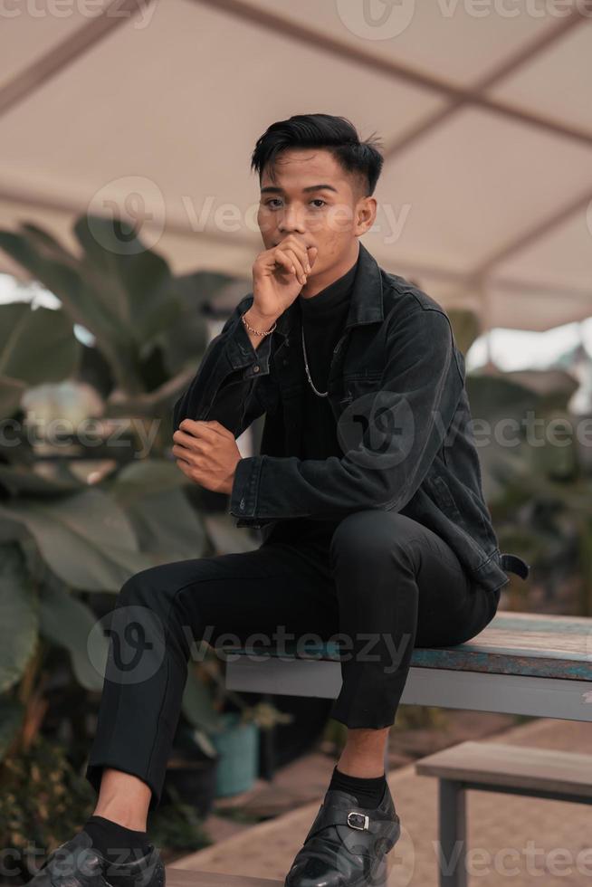 an asian boy in a black denim jacket posing like a naughty boy with a chain around his neck in a park photo