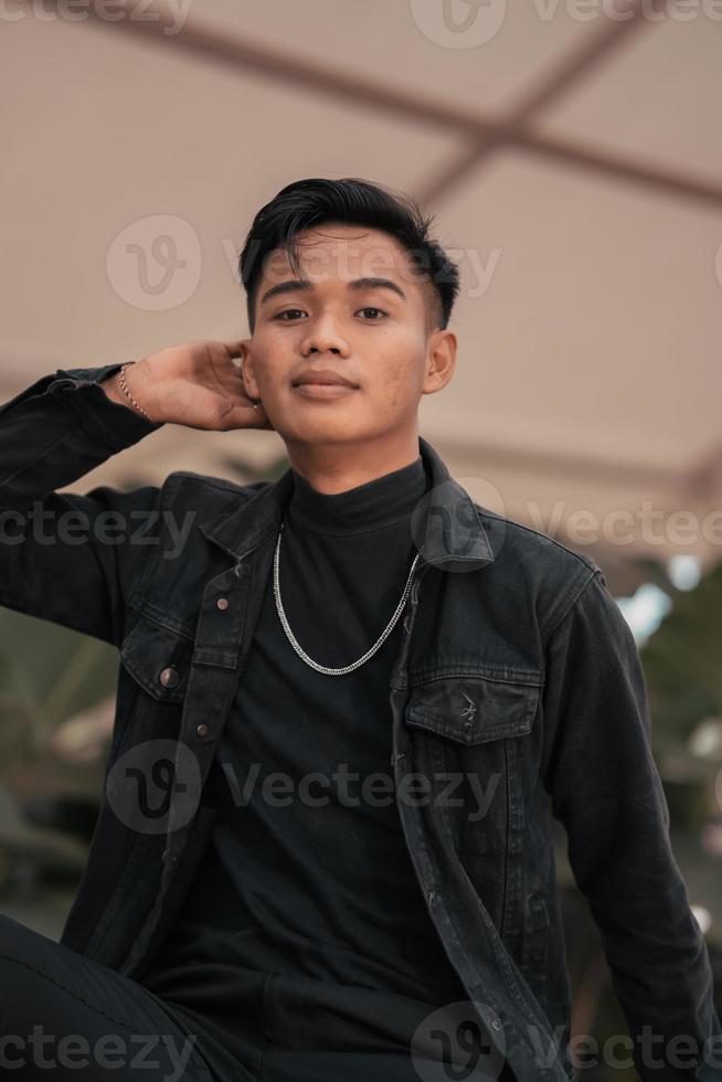 an Asian man in a black denim jacket and a chain necklace posing in a relaxed style in a park photo