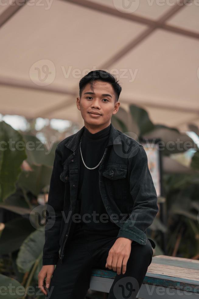 an asian boy in a black denim jacket posing like a naughty boy with a chain around his neck in a park photo