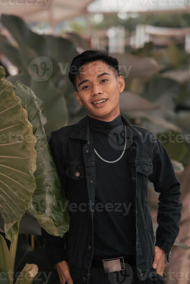 an asian boy in a black denim jacket posing like a naughty boy with a chain around his neck in a park photo