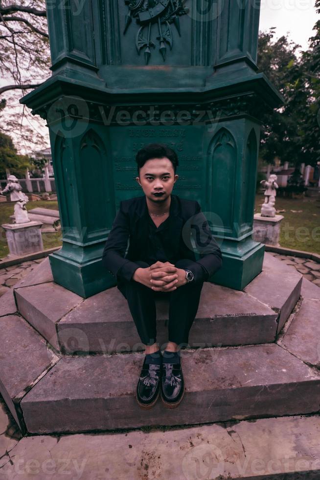 an Asian man in a black suit is sitting in a graveyard while visiting a friend who died early photo