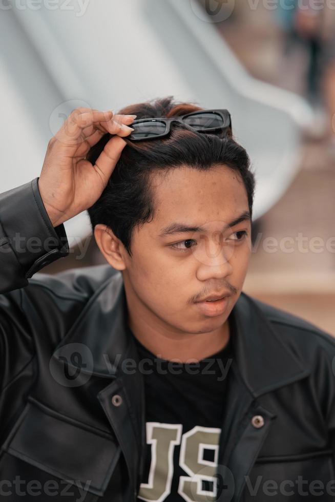 an Asian man is sitting on a wooden chair while wearing a black leather shirt and jacket while stylish with sunglasses photo