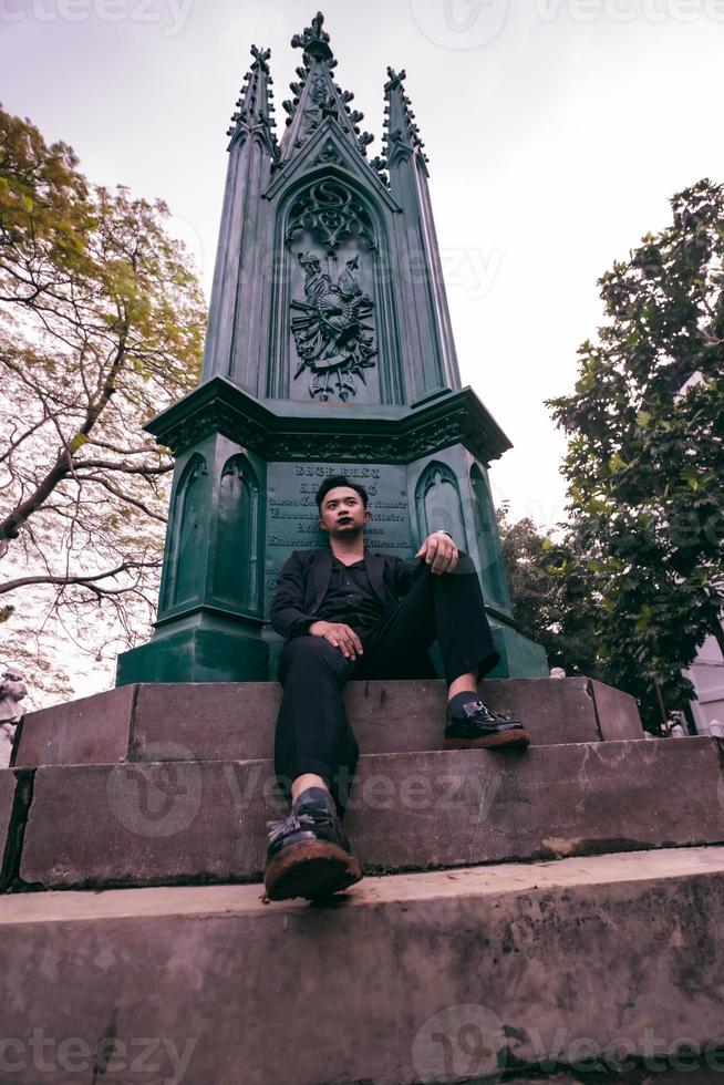 an Asian man in a black suit is sitting in a graveyard while visiting a friend who died early photo