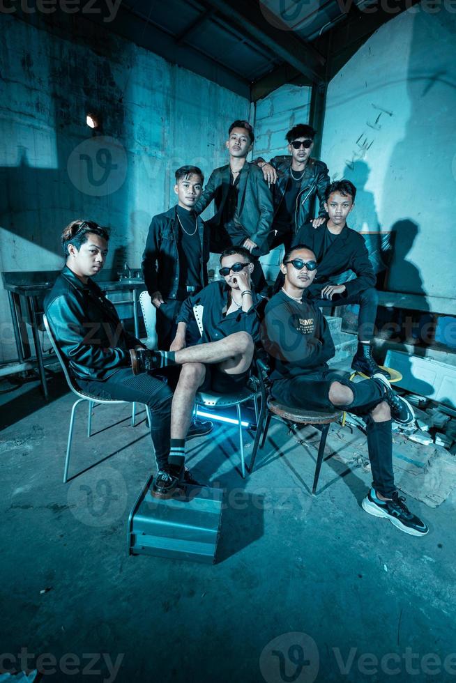 a group of Asian teenagers in black clothes posing very coolly while sitting in a warehouse chair which is lit blue photo