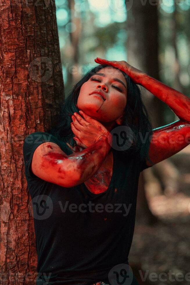 an Asian woman in black clothes smeared her body with red blood before the ritual begins in front of a big tree photo