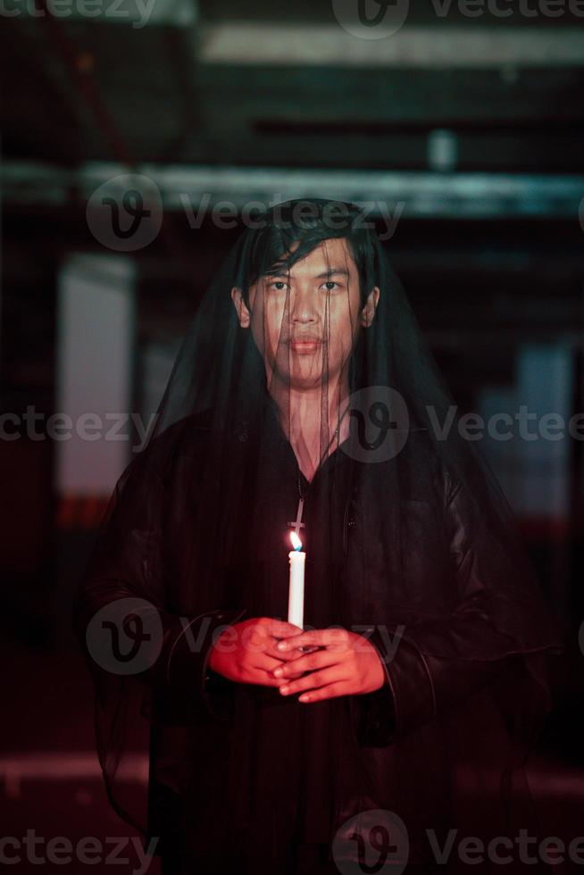 a male devil worshiper with a transparent veil is performing a spooky ritual by holding a candle in his hand photo