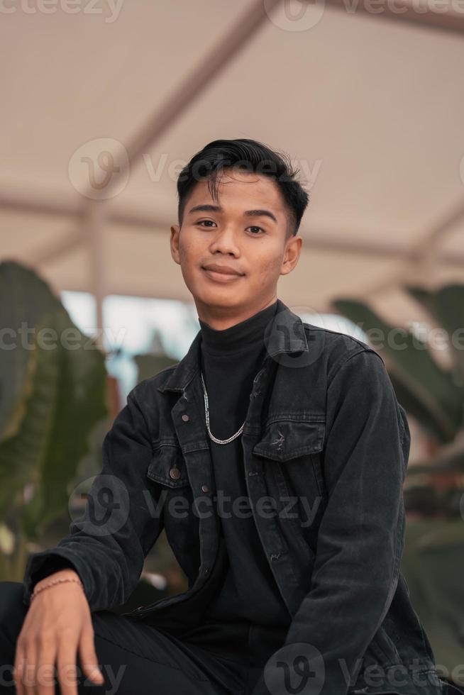 an Asian man in a black denim jacket and a chain necklace posing in a relaxed style in a park photo