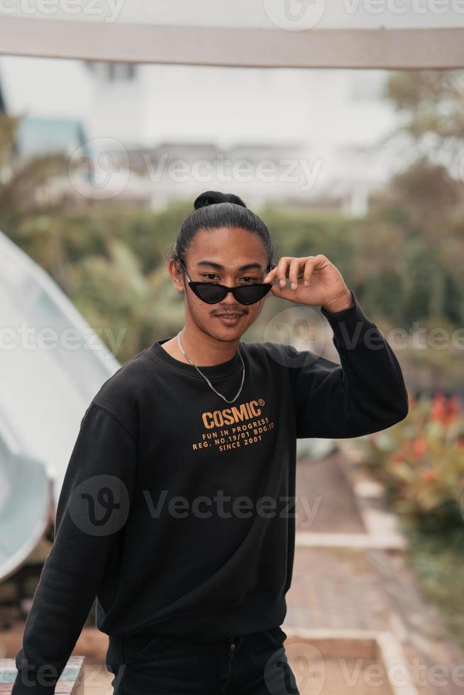 an Asian teenager in a shirt and sunglasses stands very masculine while posing in a park photo