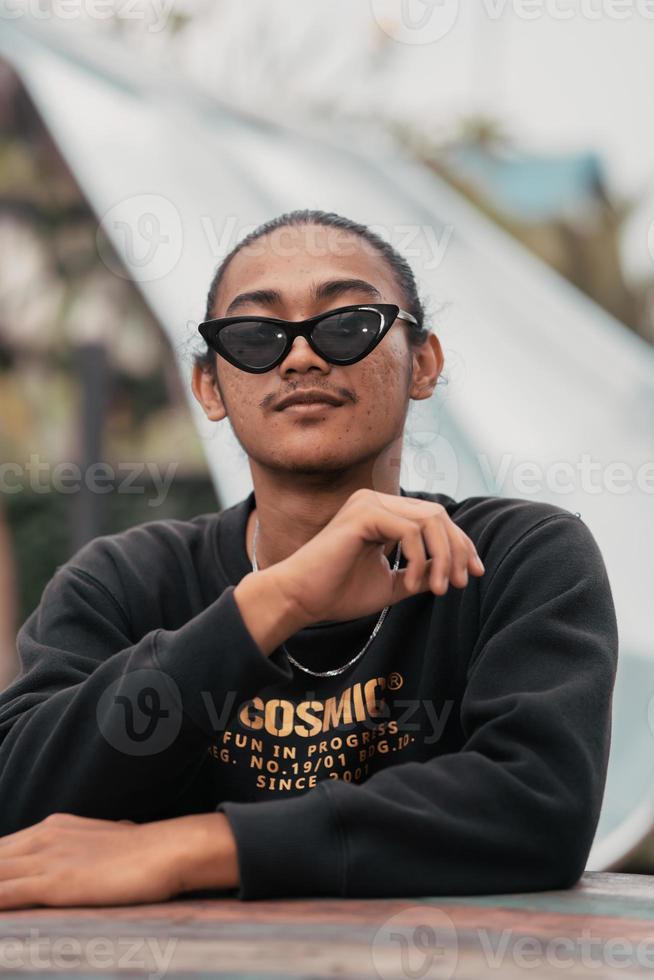 an Asian man with his hair tied up and wearing a black sweater jacket is sitting relaxing at a