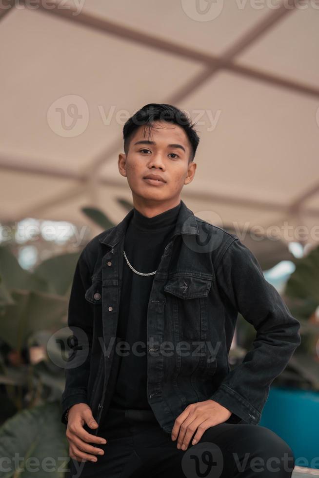 an Asian man in a black denim jacket and a chain necklace posing in a relaxed style in a park photo