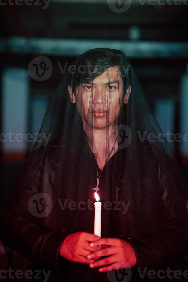 a male devil worshiper with a transparent veil is performing a spooky ritual by holding a candle in his hand photo