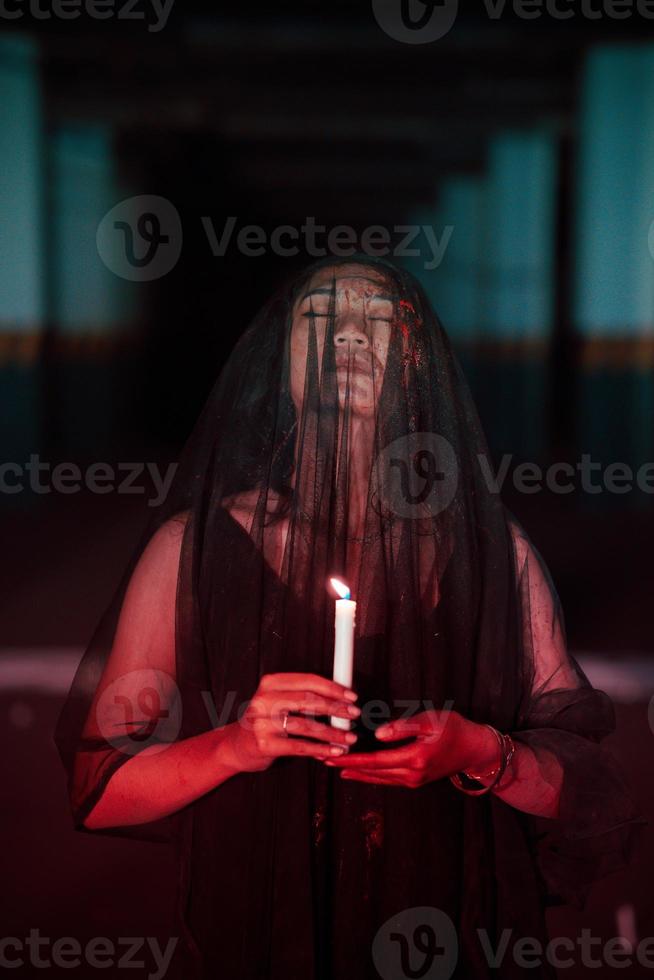 an Asian woman with closed eyes and a transparent veil stained with blood holds a candle in her hand during satanic worship photo