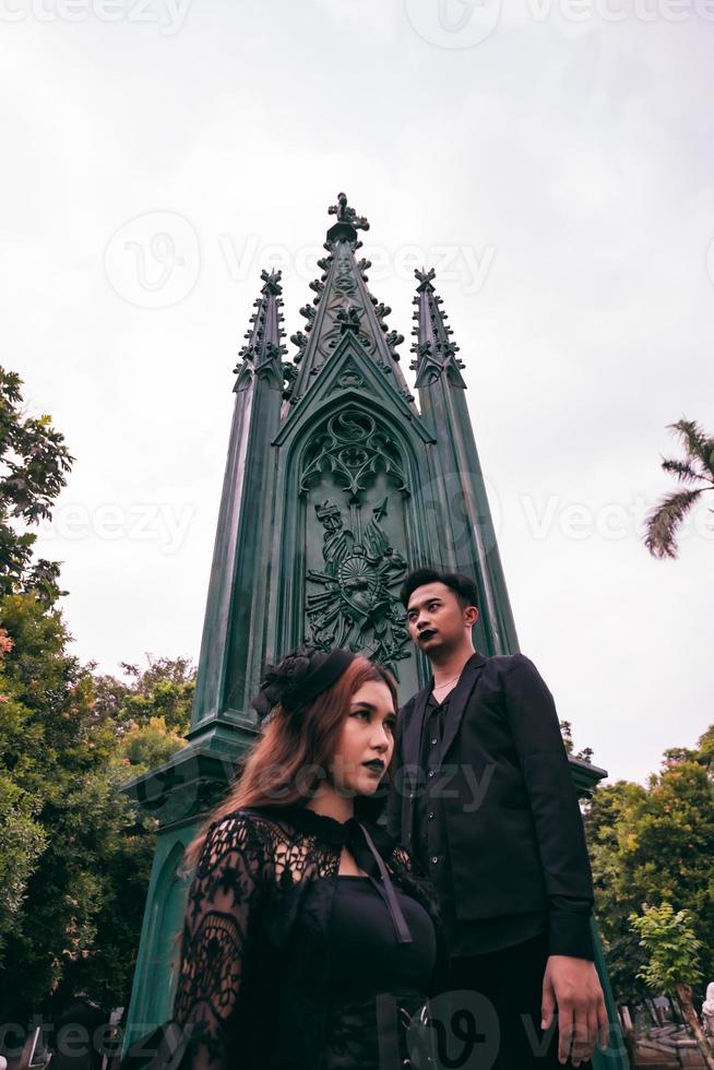 a couple of Asian teens standing near a spooky cemetery full of green trees photo