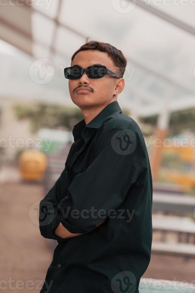 un asiático hombre con un Bigote cara vistiendo un camisa y Gafas de sol mientras colgando fuera con su amigos en un café foto