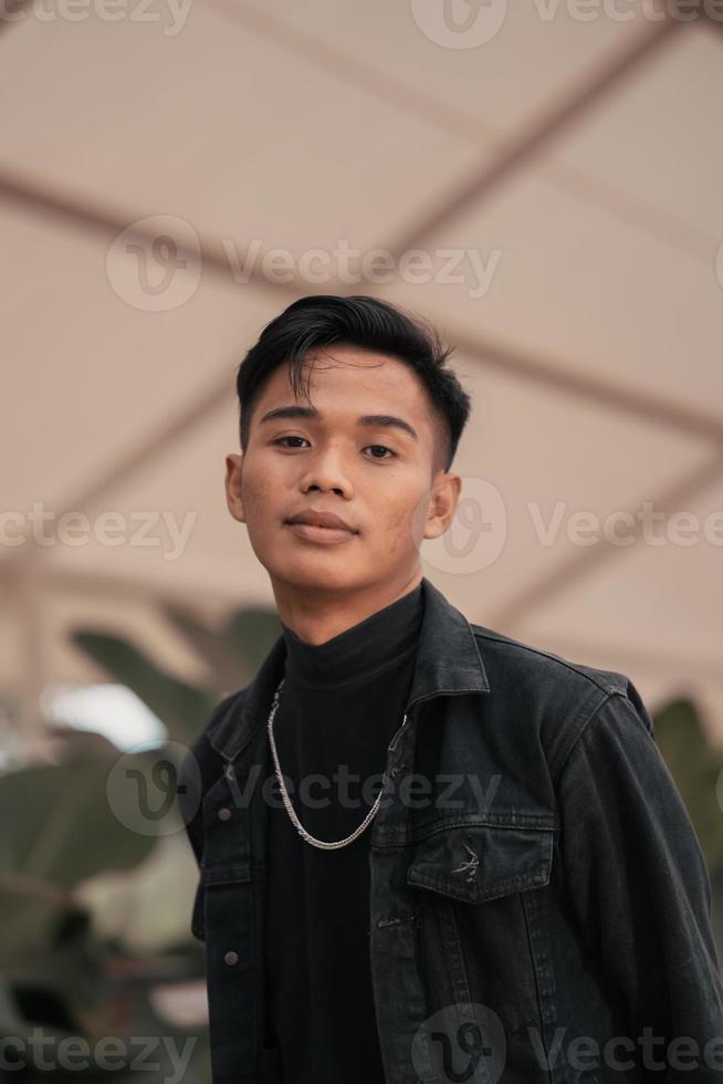 an asian boy in a black denim jacket posing like a naughty boy with a chain around his neck in a park photo