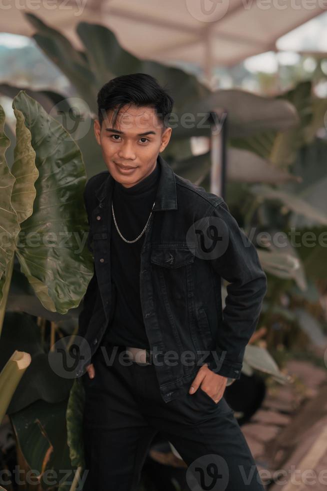 un asiático chico en un negro mezclilla chaqueta posando me gusta un travieso chico con un cadena alrededor su cuello en un parque foto