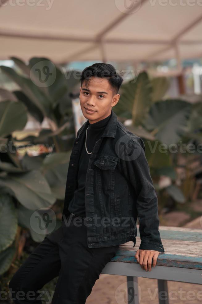 un asiático chico en un negro mezclilla chaqueta posando me gusta un travieso chico con un cadena alrededor su cuello en un parque foto