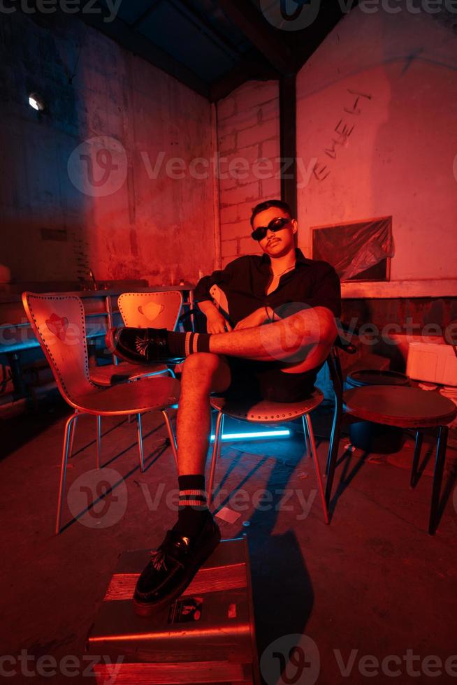 an Asian man with sunglasses sitting posing on a warehouse chair very sensually accompanied by a red light photo