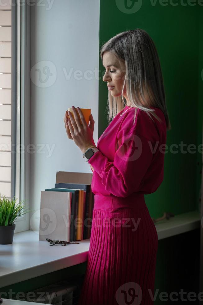 una hermosa joven rubia con un brillante traje rosa-rojo se encuentra cerca de la ventana de su oficina, descansa y bebe café caliente de una taza. foto