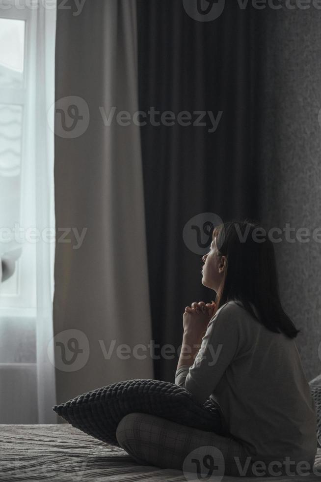 A sad young woman in pajamas with seasonal affective disorder or depression sits alone on the bed and looks out the window. The concept of winter depression due to lack of sunlight photo