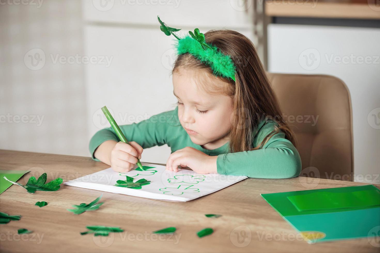 A little girl with a shamrock headband draws and cuts green shamrocks for St. Patrick's Day at her table at home photo