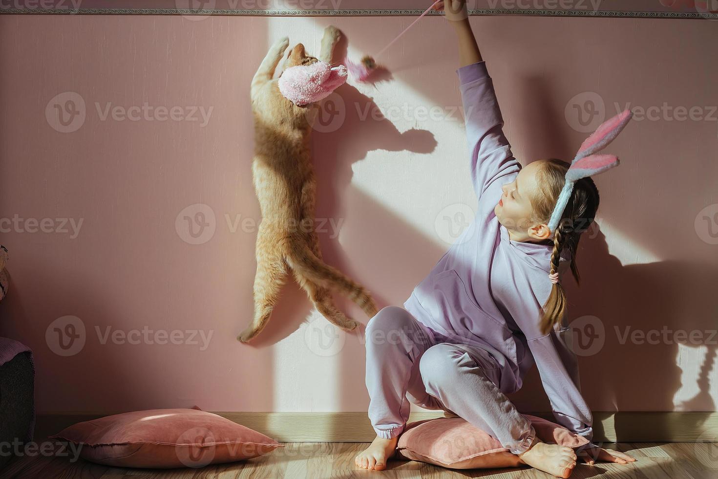 a schoolgirl girl on her head a headband with bunny ears plays with a red domestic cat in an Easter hat in the sun photo