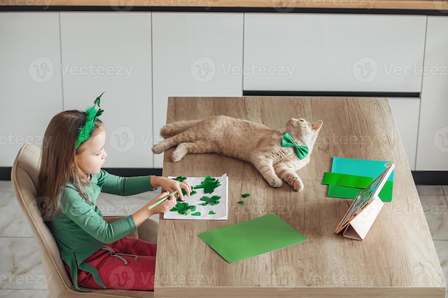 A little girl with a bandage on her head draws and cuts green shamrocks for St. Patrick's Day at a table at home in the kitchen, next to her is her beautiful cat with a green bow tie around his neck photo