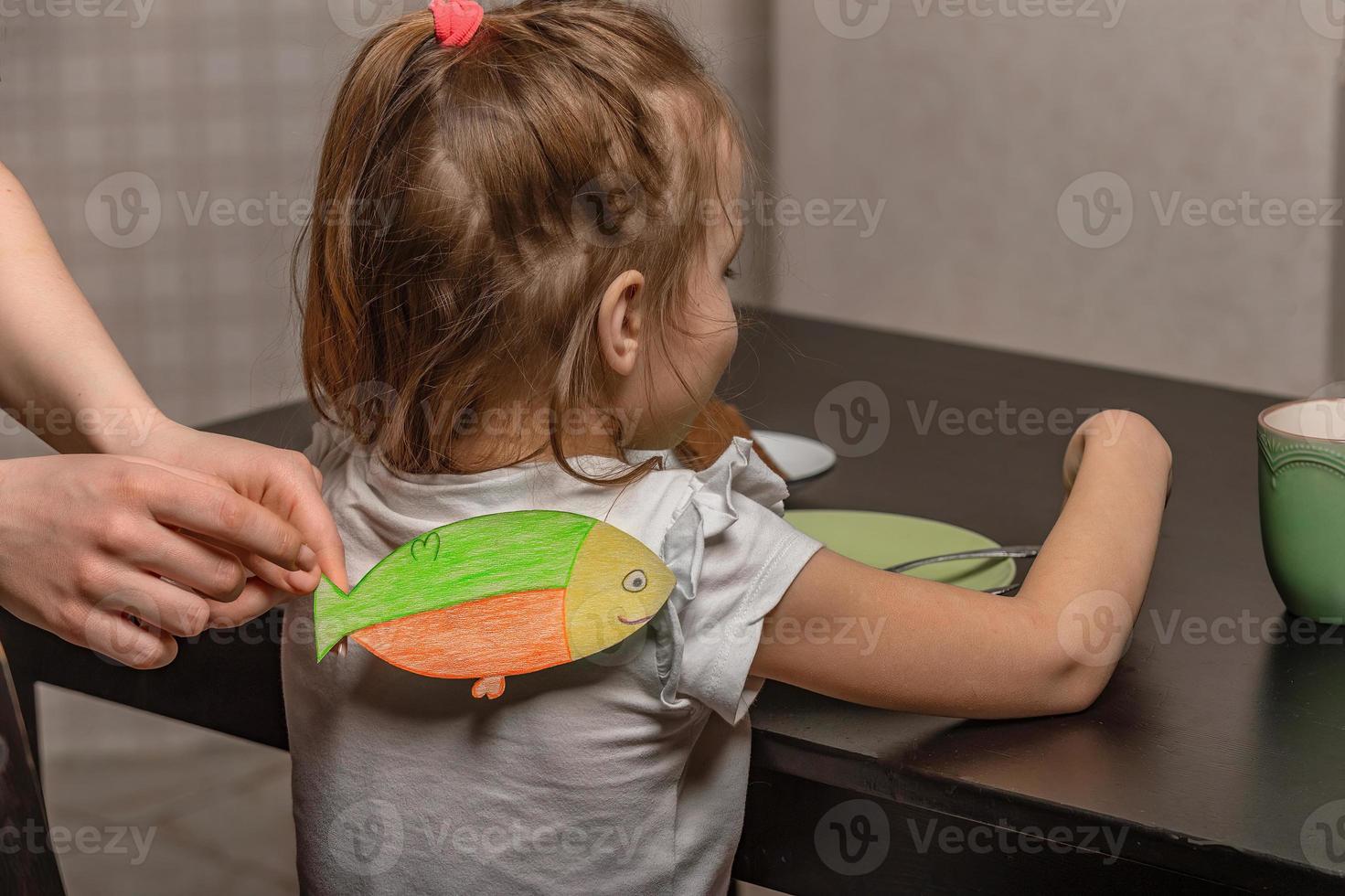 The older sister glues a paper fish to her little sister on her back at home in the kitchen, the child has breakfast. Funny family jokes, April Fool's Day celebration photo
