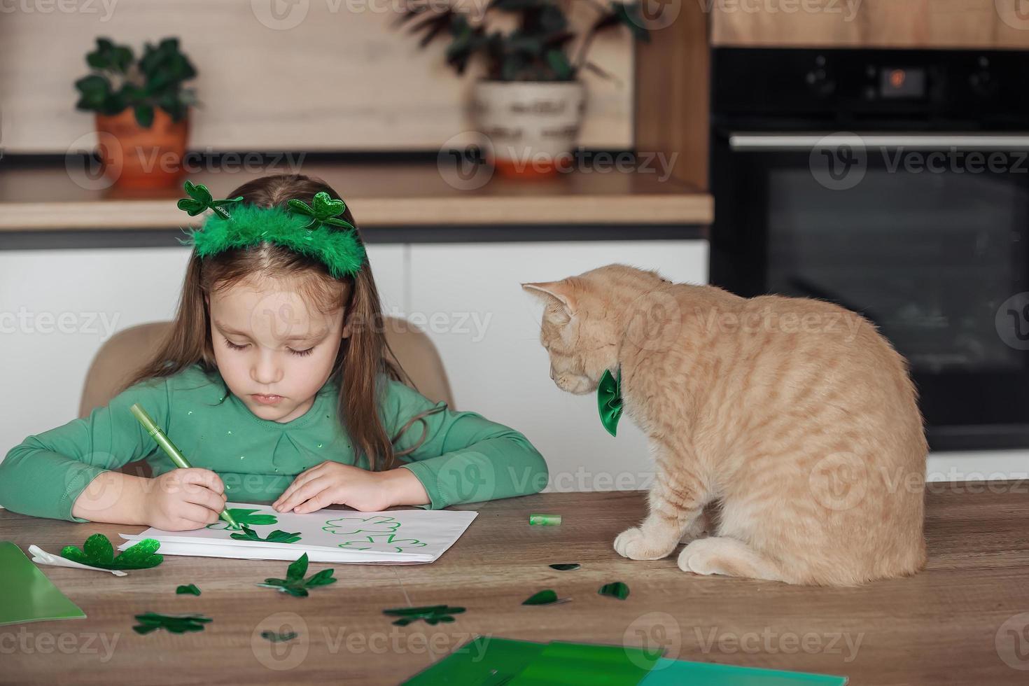 A little girl with a bandage on her head draws and cuts green shamrocks for St. Patrick's Day at a table at home in the kitchen, next to her is her beautiful cat with a green bow tie around his neck photo