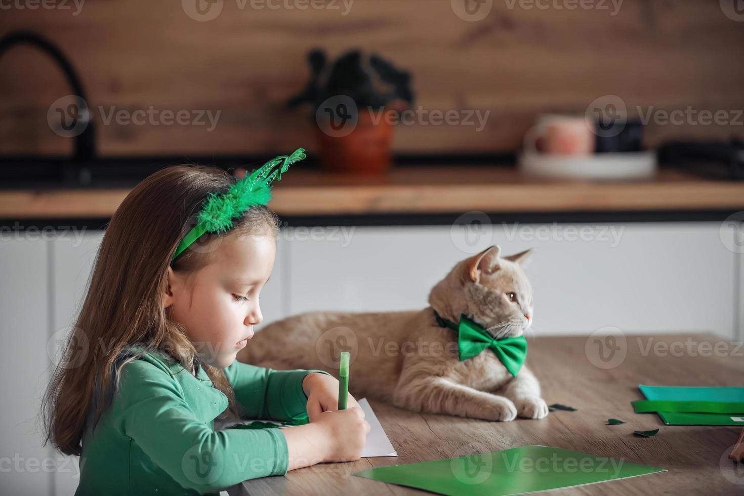 A little girl with a bandage on her head draws and cuts green shamrocks for St. Patrick's Day at a table at home in the kitchen, next to her is her beautiful cat with a green bow tie around his neck photo