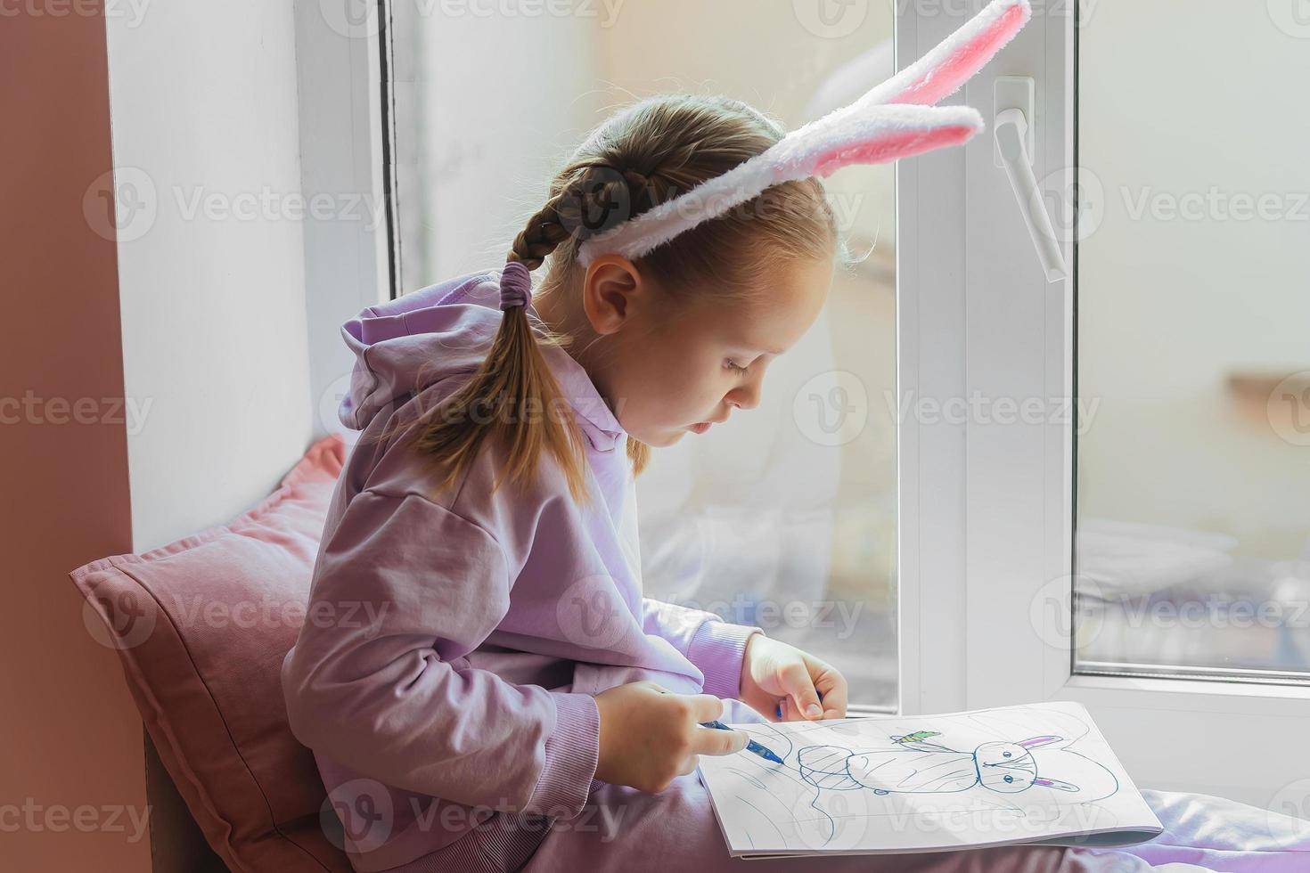a schoolgirl girl with rabbit ears sits on the window and draws a rabbit in her album, preparing for Easter photo