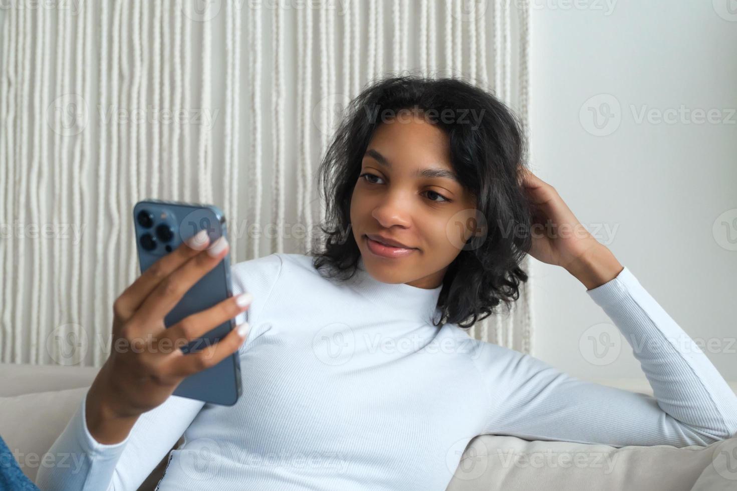 Close up view of Smiling afro american millennial woman user with smart phone watches video, playing game, shopping, chatting in mobile technology application sitting on sofa at home.Selective focus photo