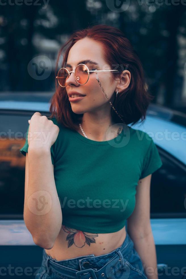 Beautiful fashionable young woman in glasses posing near car photo