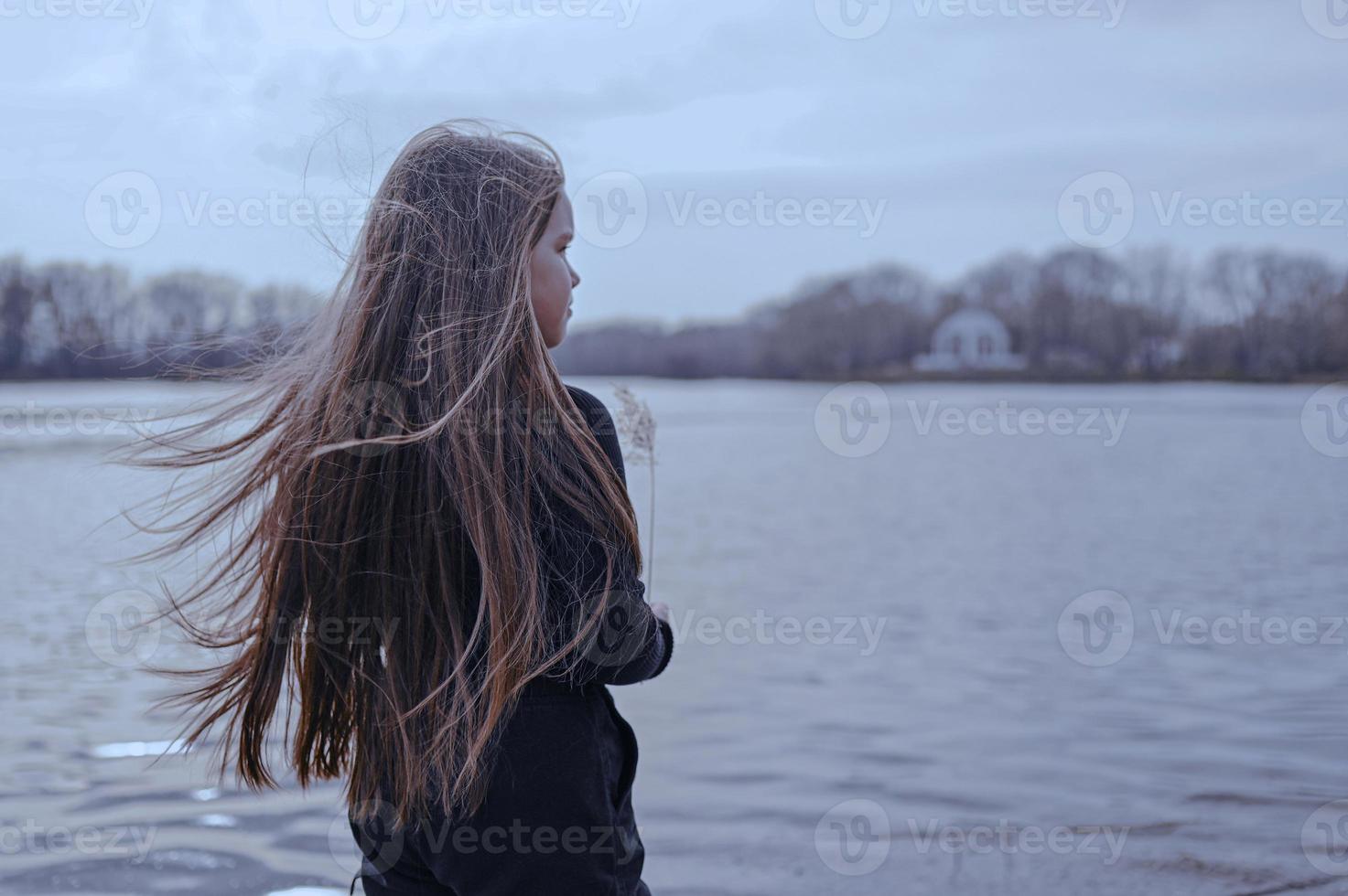 un niña con largo pelo en el apuntalar de un lago. en pie en el viento y mirando a un hermosa calentar puesta de sol. foto