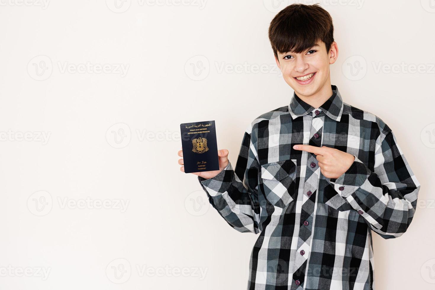 joven adolescente chico participación Siria pasaporte mirando positivo y contento en pie y sonriente con un confidente sonrisa en contra blanco antecedentes. foto
