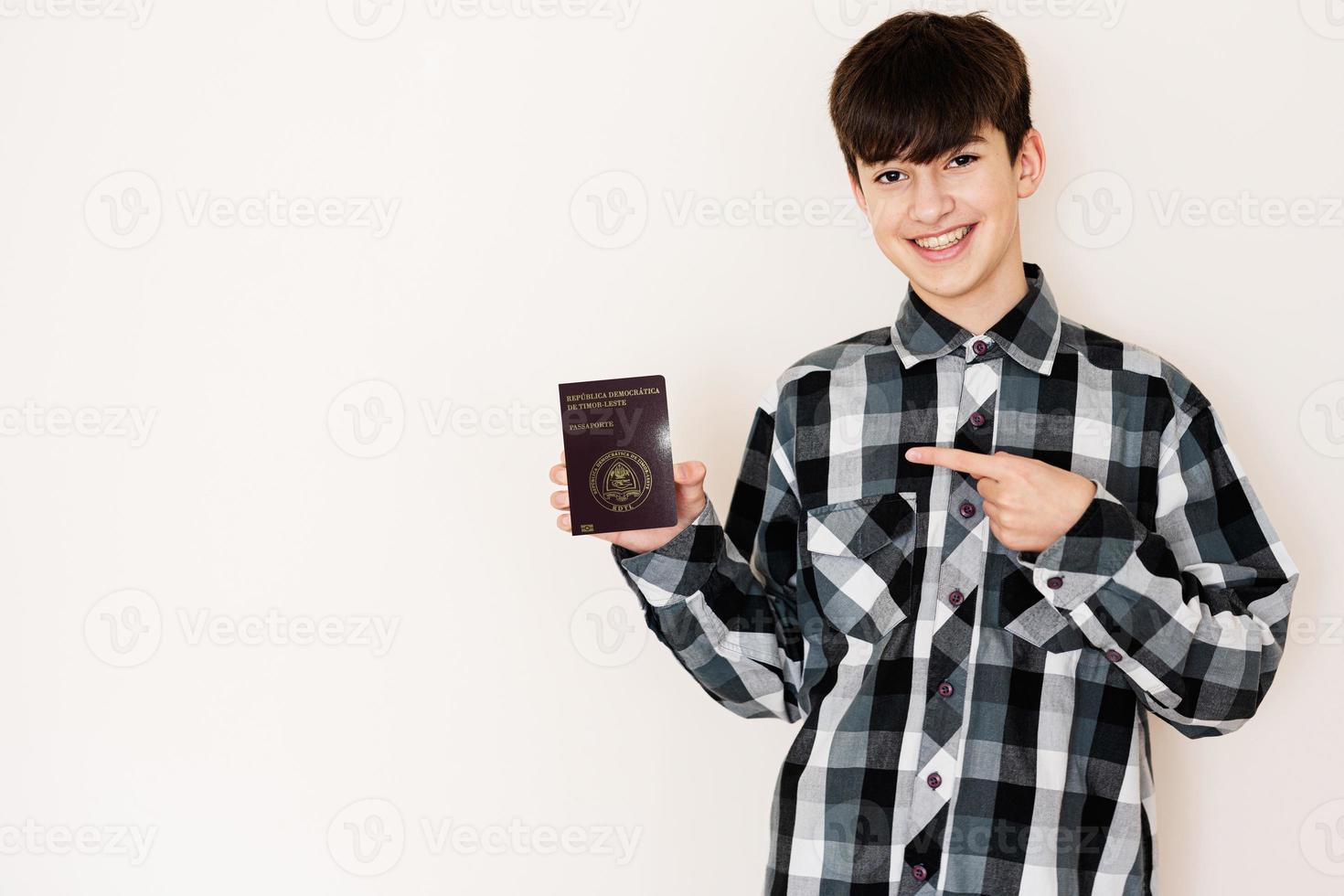 joven adolescente chico participación este Timor pasaporte mirando positivo y contento en pie y sonriente con un confidente sonrisa en contra blanco antecedentes. foto