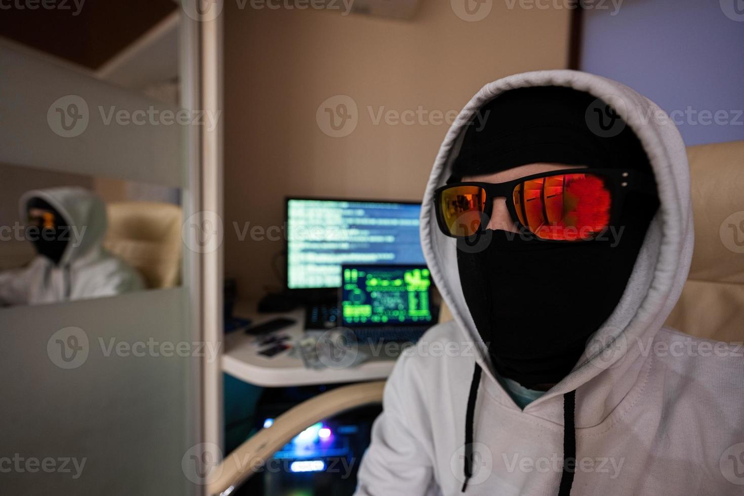 Close up of boy hacker. Internet theft . Man wearing a balaclava and sunglasses sat behind a laptop. photo