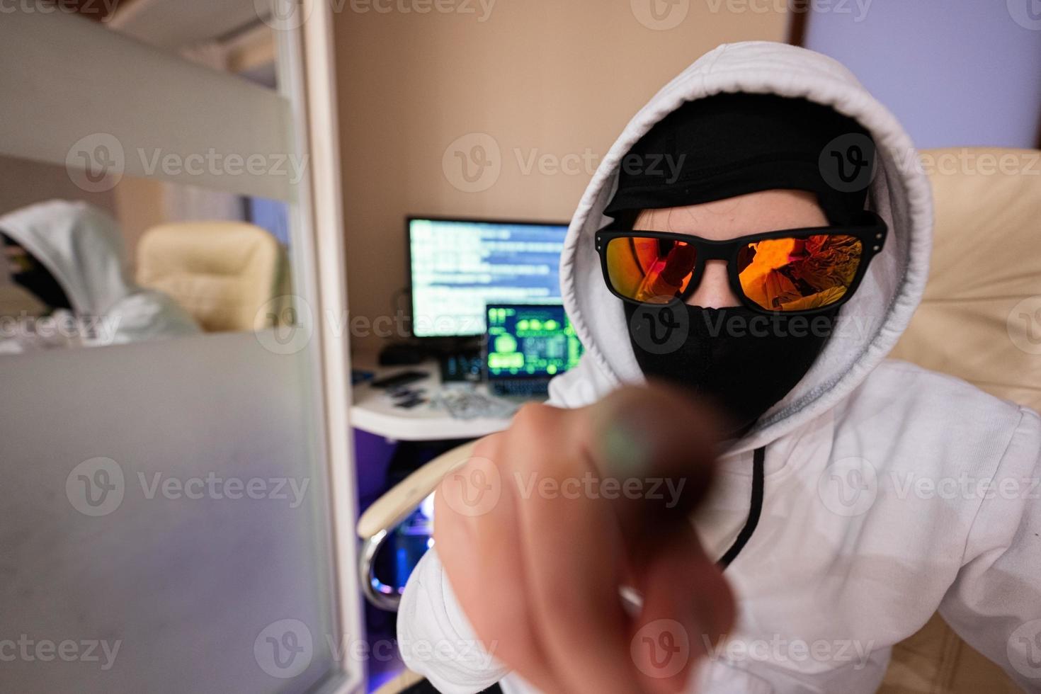 Close up of boy hacker. Internet theft show finger to screen . Man wearing a balaclava and sunglasses sat behind a laptop. photo