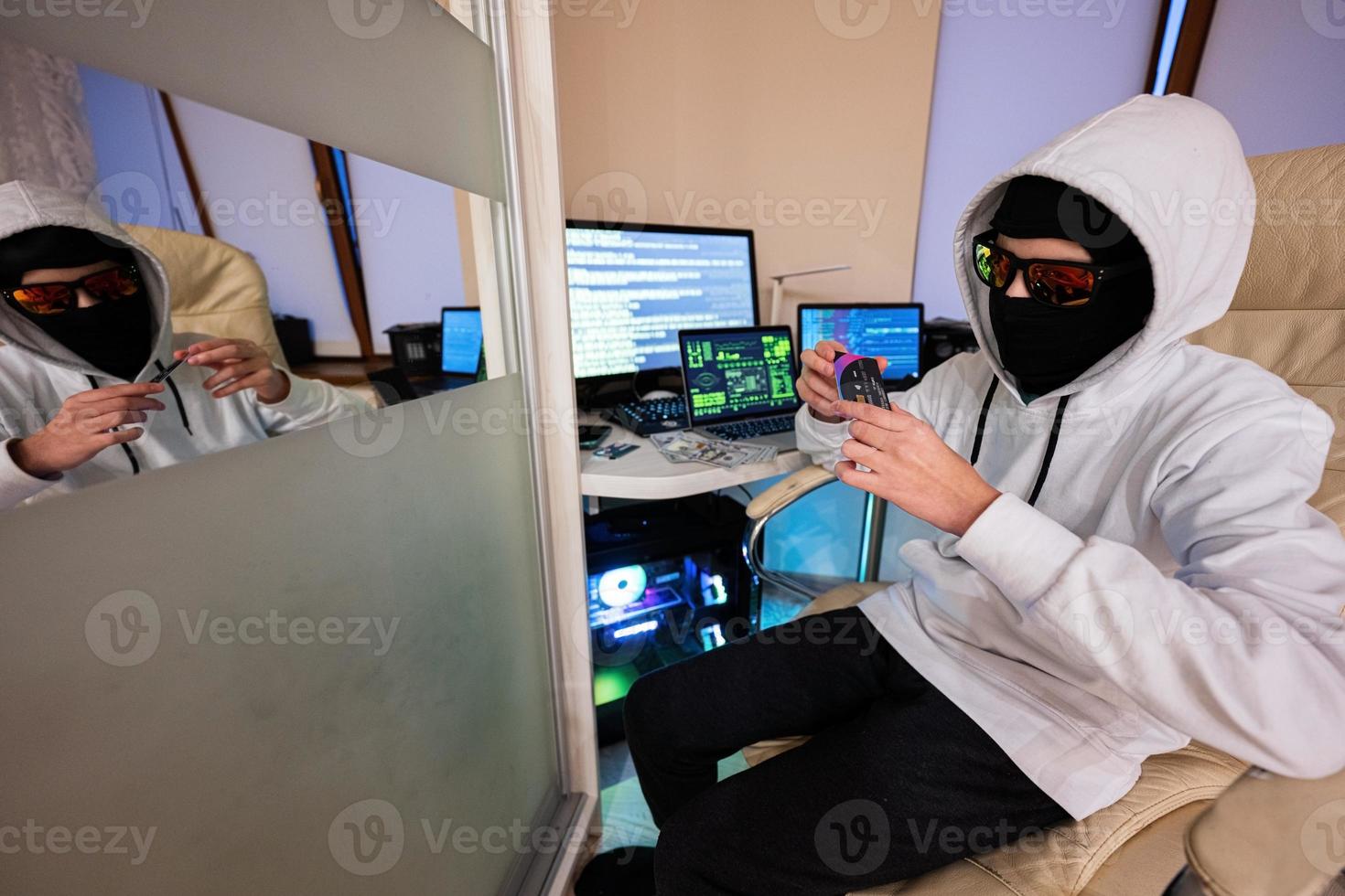 Boy hacker fraudulently use credit card for payment. Internet theft . Man wearing a balaclava and holding a credit card while sat behind a laptop. photo