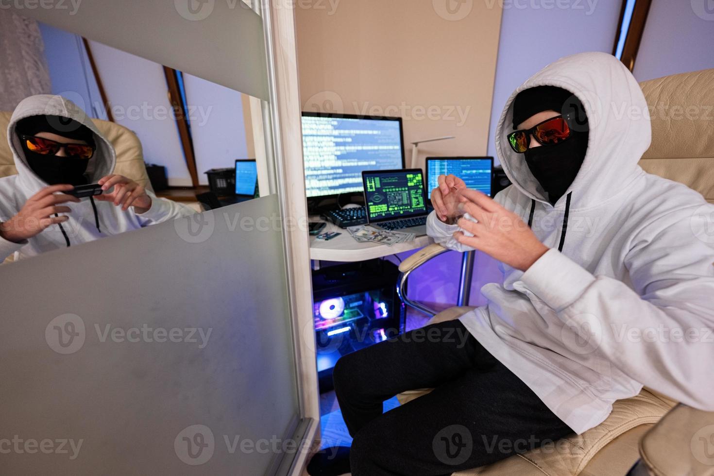 Boy hacker fraudulently use credit card for payment. Internet theft . Man wearing a balaclava and holding a credit card while sat behind a laptop. photo