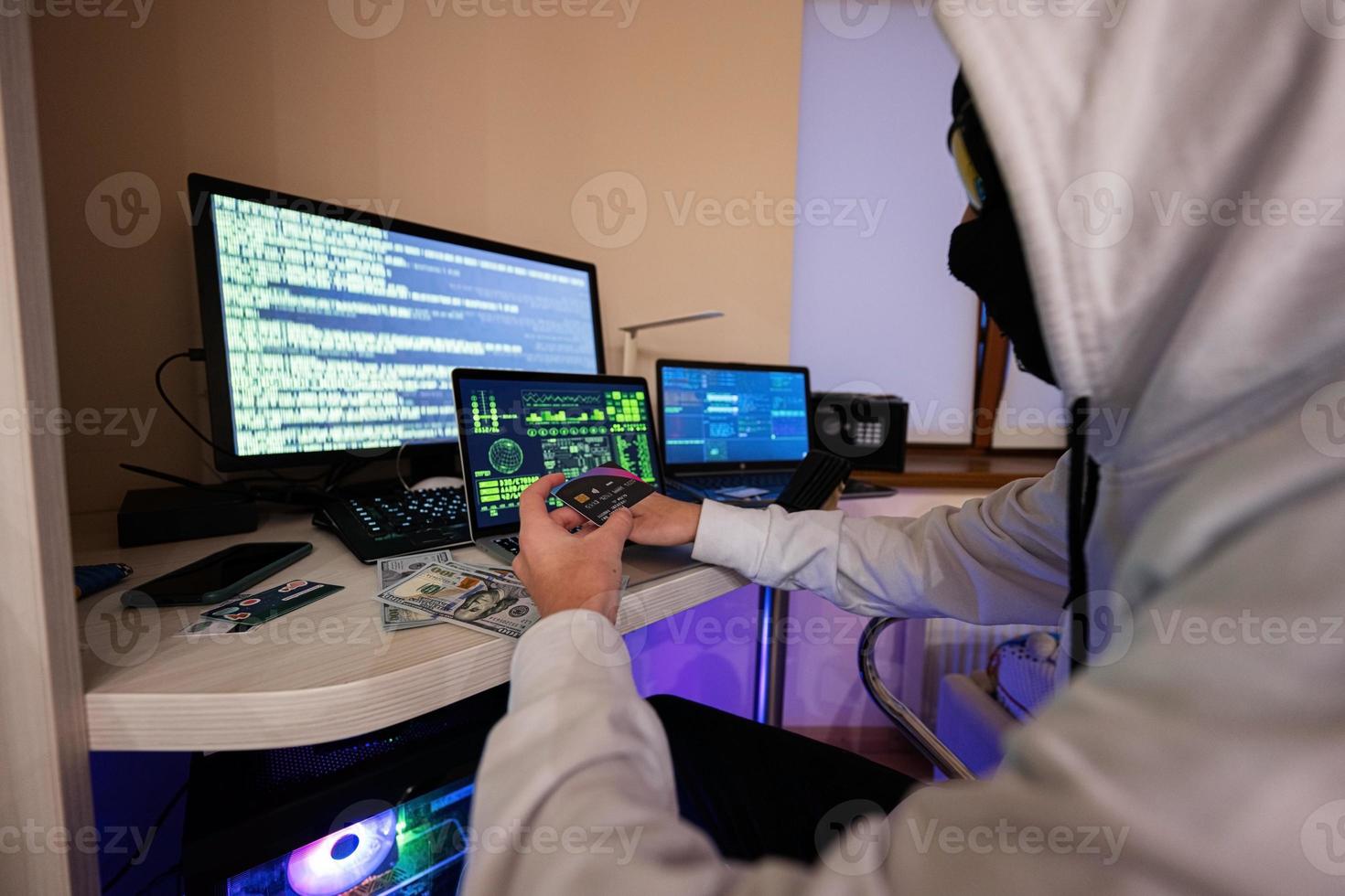 Boy hacker fraudulently use credit card for payment. Internet theft . Man wearing a balaclava and holding a credit card while sat behind a laptop. photo