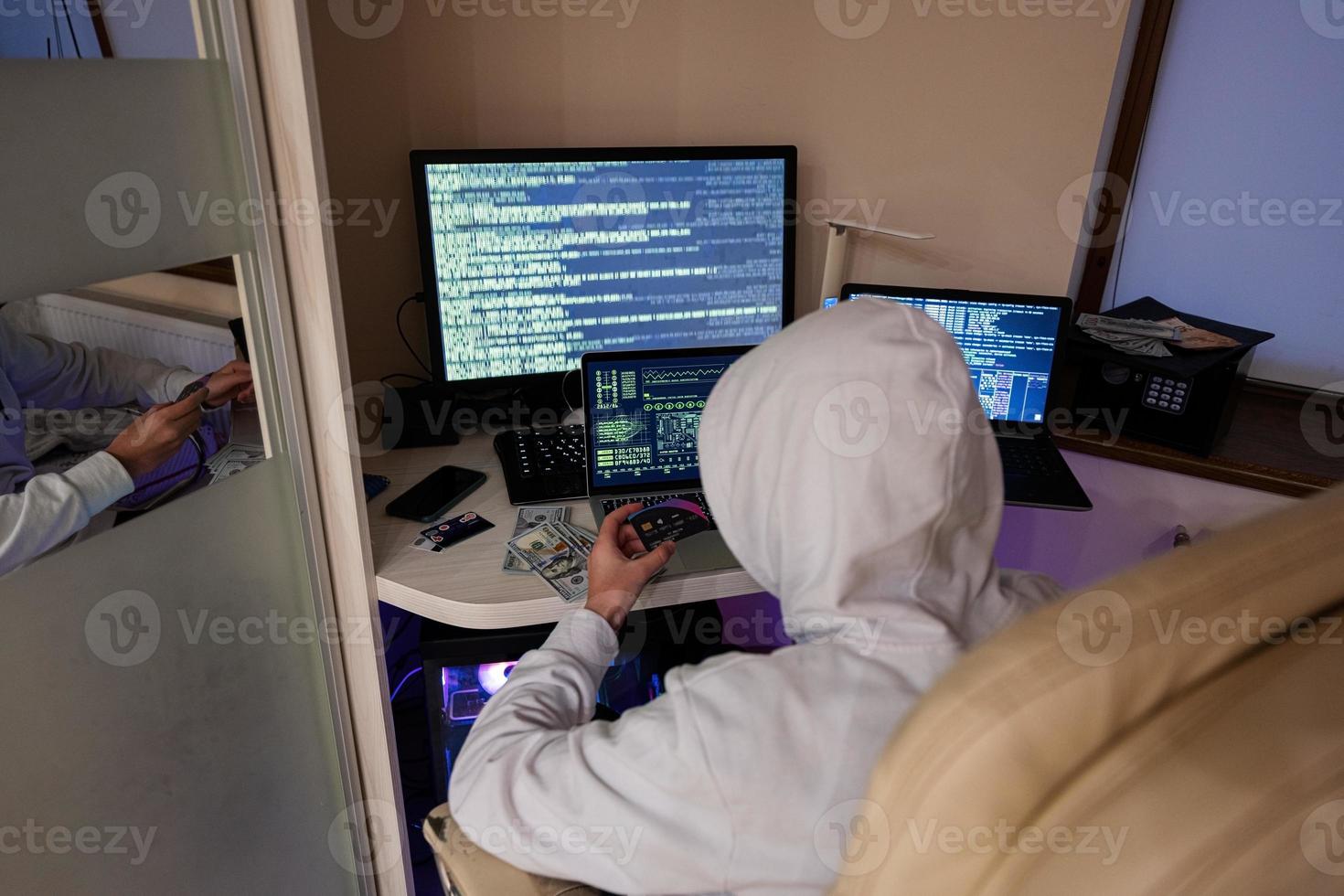 Boy hacker fraudulently use credit card for payment. Internet theft . Man wearing a balaclava and holding a credit card while sat behind a laptop. photo