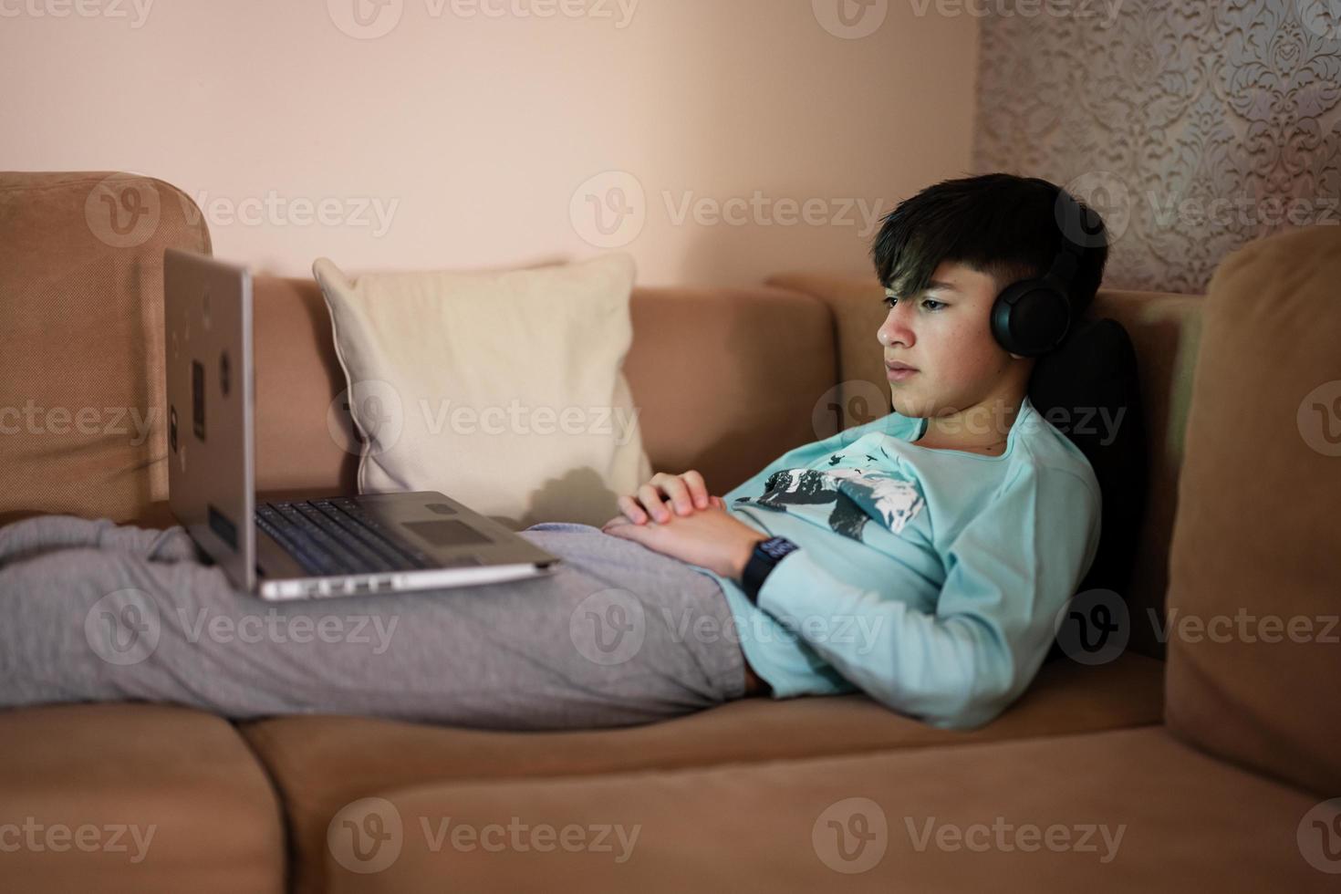 joven adolescente chico con auriculares en frente de un ordenador portátil en un cama a noche. foto