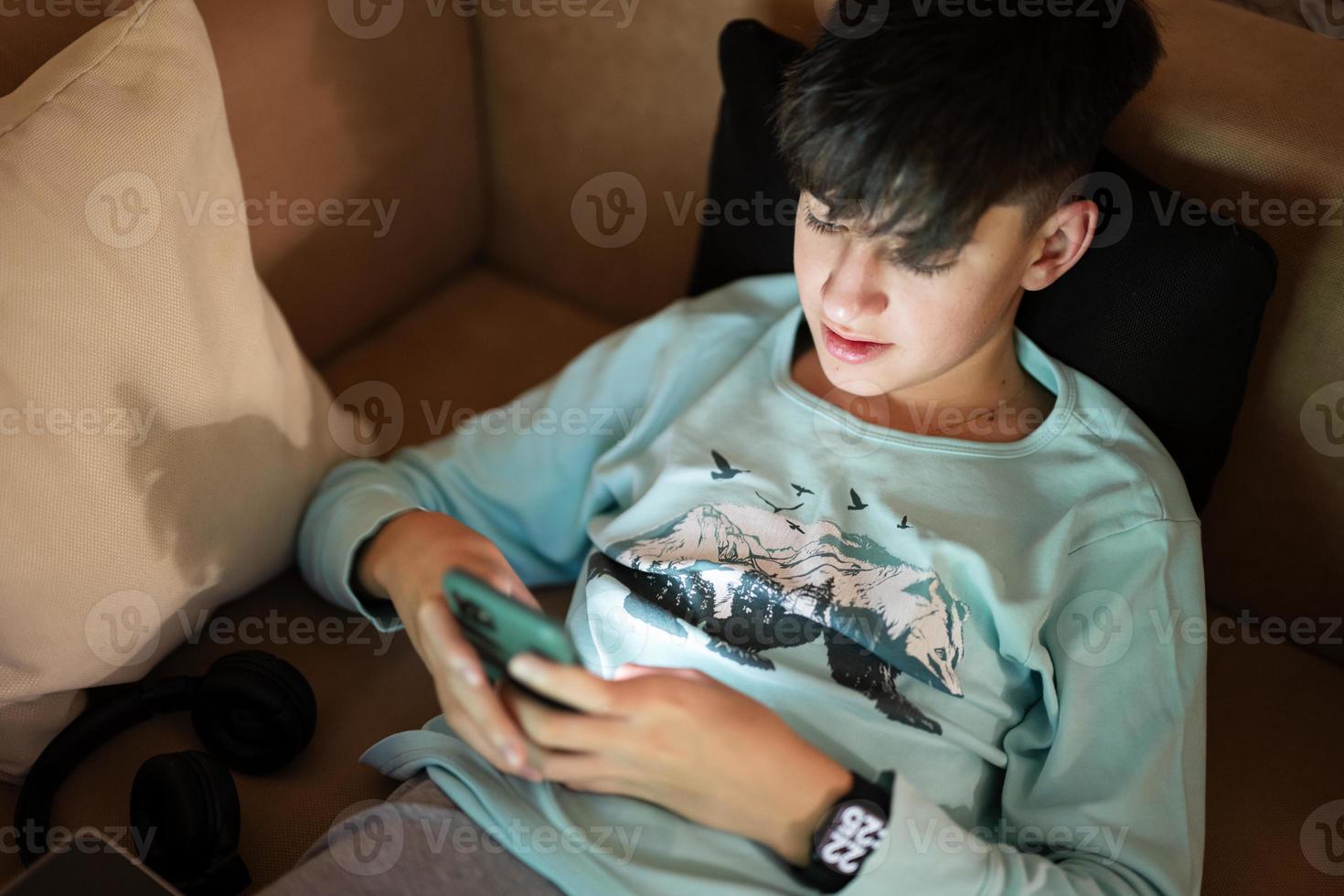 joven adolescente chico mensajes de texto teléfono en frente de un ordenador portátil en un cama a noche. foto