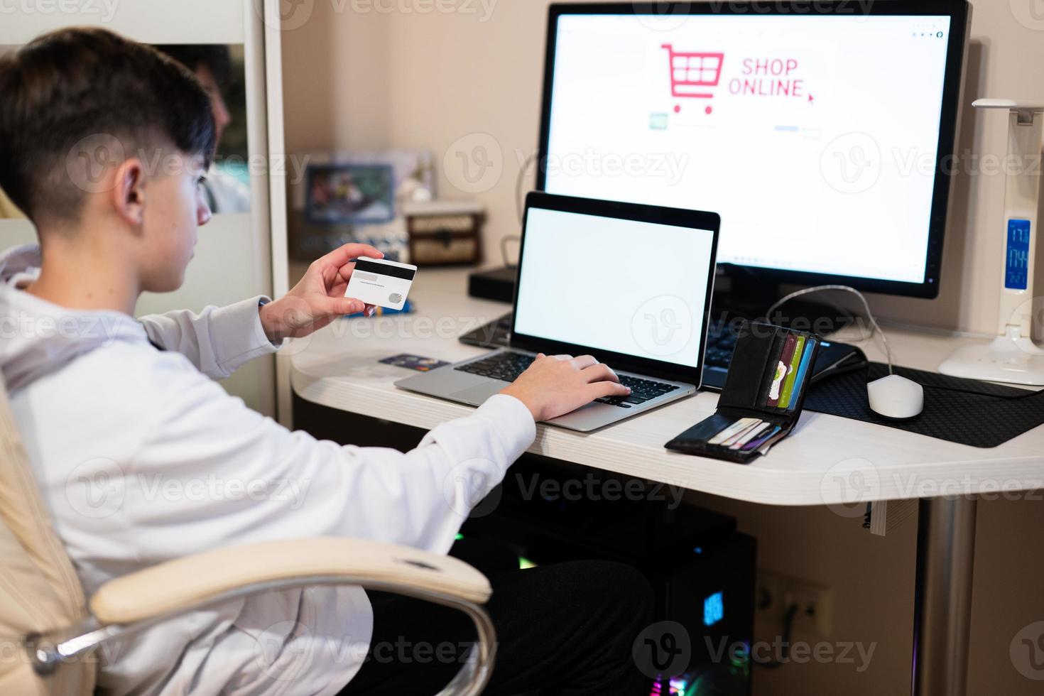 Happy teenage boy using laptop and credit card, sitting at desk and ordering goods online from home. photo