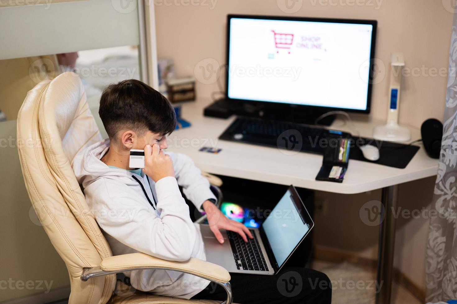 Happy teenage boy using laptop and credit card, sitting at desk and ordering goods online from home. photo