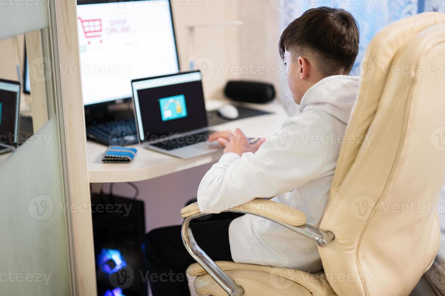 Happy teenage boy using laptop and credit card, sitting at desk and ordering goods online from home. photo